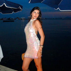 A woman posing in a shimmering, iridescent halter mini dress at a waterfront during twilight, with blue umbrellas and water in the background.