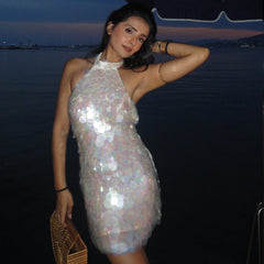 A woman posing outdoors at dusk under a blue patio umbrella, wearing a shimmering silver halter mini dress and holding a woven clutch bag.