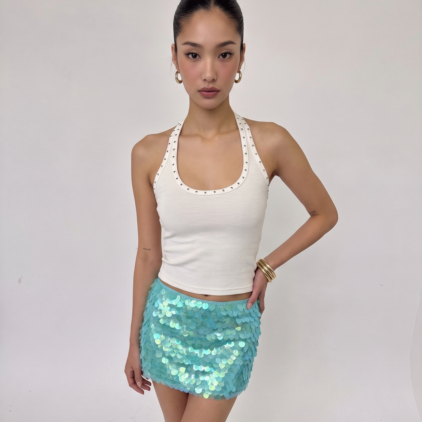 A woman modeling a white halter crop top with scalloped edges and a light blue sequined mini skirt, standing against a plain light gray studio background.