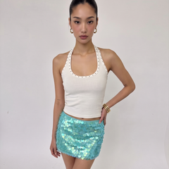 A woman modeling a white halter crop top with scalloped edges and a light blue sequined mini skirt, standing against a plain light gray studio background.