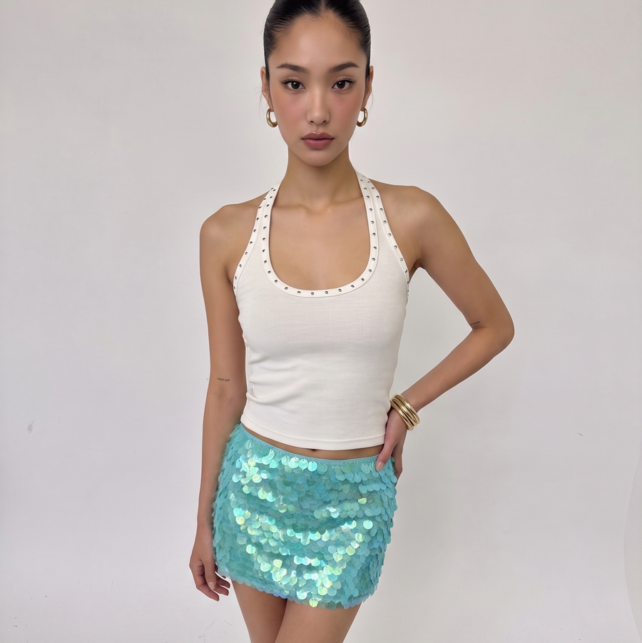 A woman modeling a white halter crop top with scalloped edges and a light blue sequined mini skirt, standing against a plain light gray studio background.