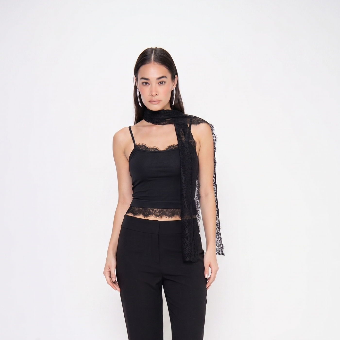 A model stands against a plain white studio background wearing a black lace-trimmed camisole top with a sheer draped scarf detail, paired with black wide-leg trousers and black pointed shoes.