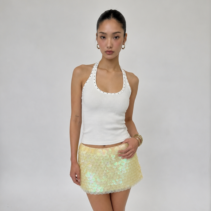 A model stands against a light gray studio background wearing a white ribbed knit halter top with scalloped trim and a short, iridescent pale yellow sequin mini skirt, paired with black flip-flop sandals.